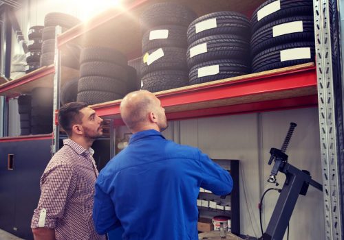 auto service, repair, maintenance and people concept - mechanic and man looking at tires at car shop